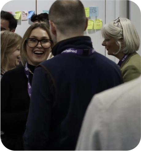 Group of attendees wearing lanyards talking at an indoor workshop; one person smiles toward the camera and a wall of colourful sticky notes is visible behind them.